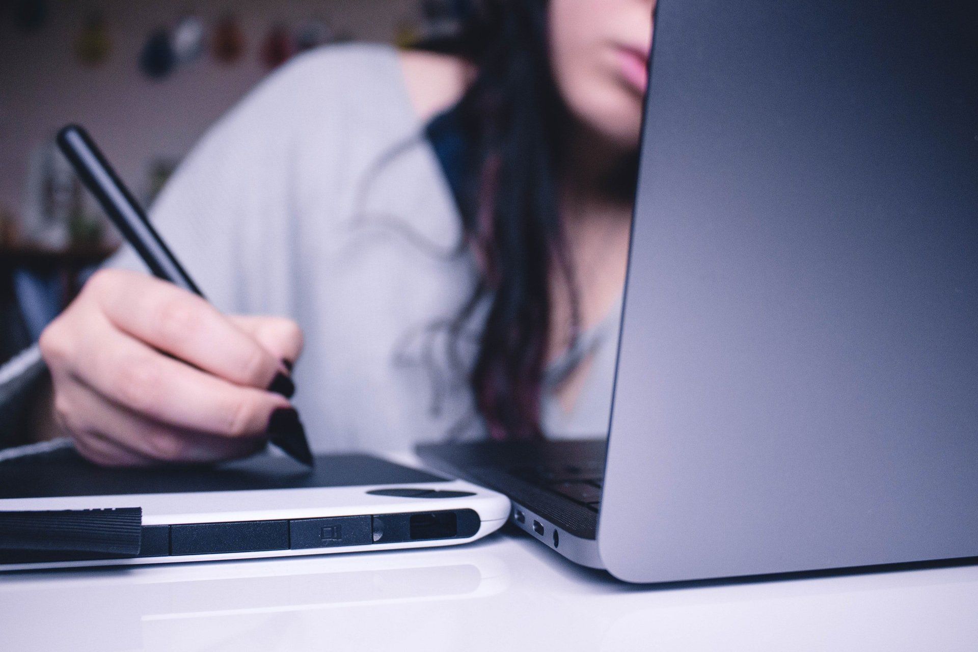 A woman is using a pen to draw on a tablet next to a laptop.